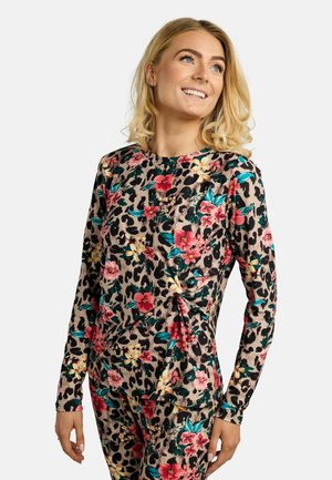 Žena s blond vlasmi sa usmieva, má na sebe zladený kvetinový a leopardí vzor na dlhom tričku a nohaviciach na jednoduchom pozadí.