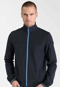 Veste bomber bleu marine foncé avec un col montant, une fermeture éclair bleue et des poignets côtelés. Comprend des poches avant et un écusson sur la manche gauche.