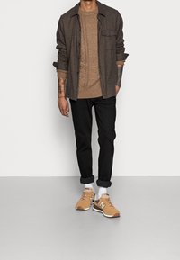 Camicia di flanella marrone su un maglione a trecce beige, pantaloni neri e sneakers beige con suole e dettagli bianchi. Outfit casual a strati.