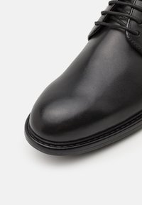 Chaussure habillée en cuir noir avec un bout arrondi, texture lisse et un laçage fin. Présente une couture discrète et une semelle en caoutchouc robuste.