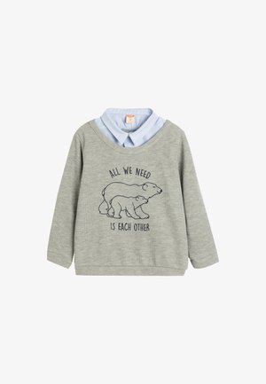 Sweatshirt gris à manches longues, avec une chemise col faux bleu clair en dessous et un motif d'ours avec le texte "Tout ce dont nous avons besoin, c'est de l'autre."