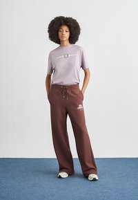 Svetlofialové tričko s logom "Nike Shox", v kombinácii s voľnými hnedými teplákmi s bielym logom "Nike" a bielymi teniskami.