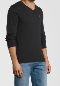 Pull noir à col en V avec des manches longues, poignets et ourlet côtelés, arborant un petit logo rouge sur la poitrine gauche. Porté sur un jean bleu clair.