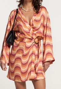 Mujer vistiendo un vestido corto de manga larga con rayas onduladas en naranja y rosa, un detalle de anillo en la cintura, collar de perlas y bolso negro de hombro.