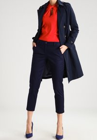 Marinblå trenchcoat med dubbelknäppning, röd blus med nyckelhålsutskärning, marinblå capribyxor och blå klackar.