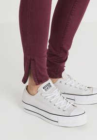 Vaqueros skinny burdeos con cremalleras en los laterales del dobladillo, combinados con zapatillas de lona blancas con acentos en negro y suela de goma.