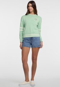 Minta zelena pulover s belimi črtastimi poudarki, rebrastimi manšetami in robom, s logotipom na prsih, v kombinaciji z jeans kratkimi hlačami in belimi športnimi copati.
