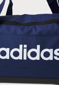 Marineblå stof duffelbag med en sort lynlås, der har et stort hvidt "adidas" logo på siden. Glat tekstur og rektangulær form.