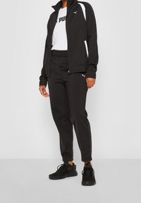 Musta urheilutakki valkoisilla yksityiskohdilla, yhdistetty mustiin housuihin ja mustiin lenkkareihin. Sisältää joustavan vyötärönauhan ja logobrändäyksen.