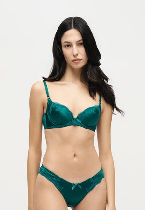 Une femme pose avec un soutien-gorge bleu canard bordé de dentelle et une culotte assortie, sur un fond clair uni.