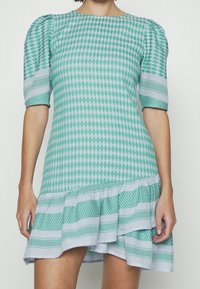 Vestido de manga corta en azul-verde con patrones de diamantes y rayas. Presenta un dobladillo con volantes y tejido texturizado contrastante en las mangas.