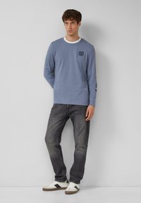 Lichtblauw langslavend T-shirt met een ronde hals, logo op de borst, gecombineerd met donkere jeans en witte sneakers. Eenvoudig, casual ontwerp.