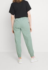 Person som bär ljusgröna byxor med elastiska ärmslut, svart kortärmad skjorta och vita sneakers, stående mot en enfärgad vit bakgrund.