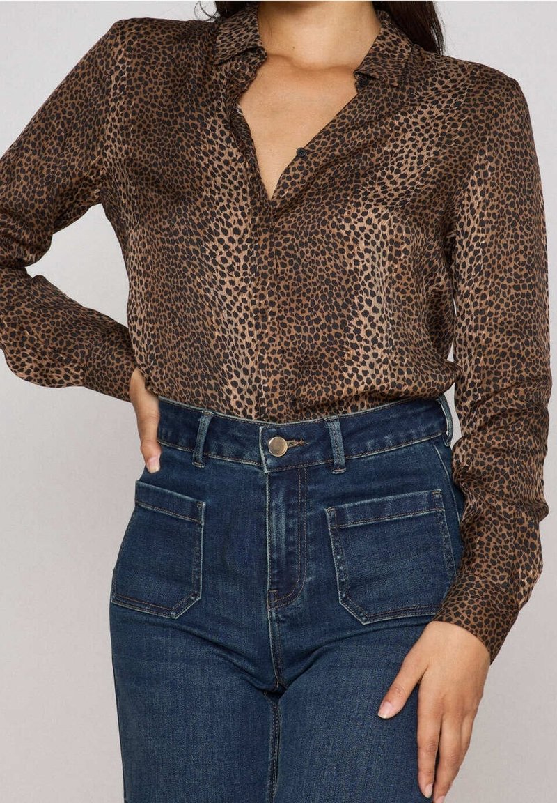 Femme portant un chemisier marron et noir à manches longues à pois, rentré dans un jean bleu foncé taille haute avec des poches plaquées à l'avant.