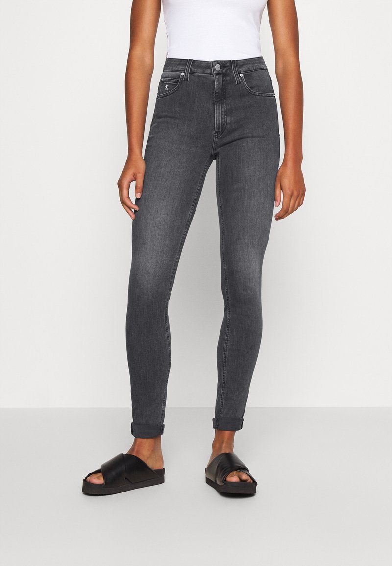 Pantalones skinny negros de cintura alta con una textura suave, diseño clásico de cinco bolsillos y puños enrollados, combinados con sandalias deslizadas negras.