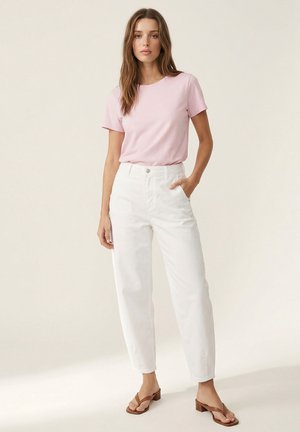 Woman standing wearing a light pink short-sleeve shirt, high-waisted white pants, and brown heeled sandals against a plain background.