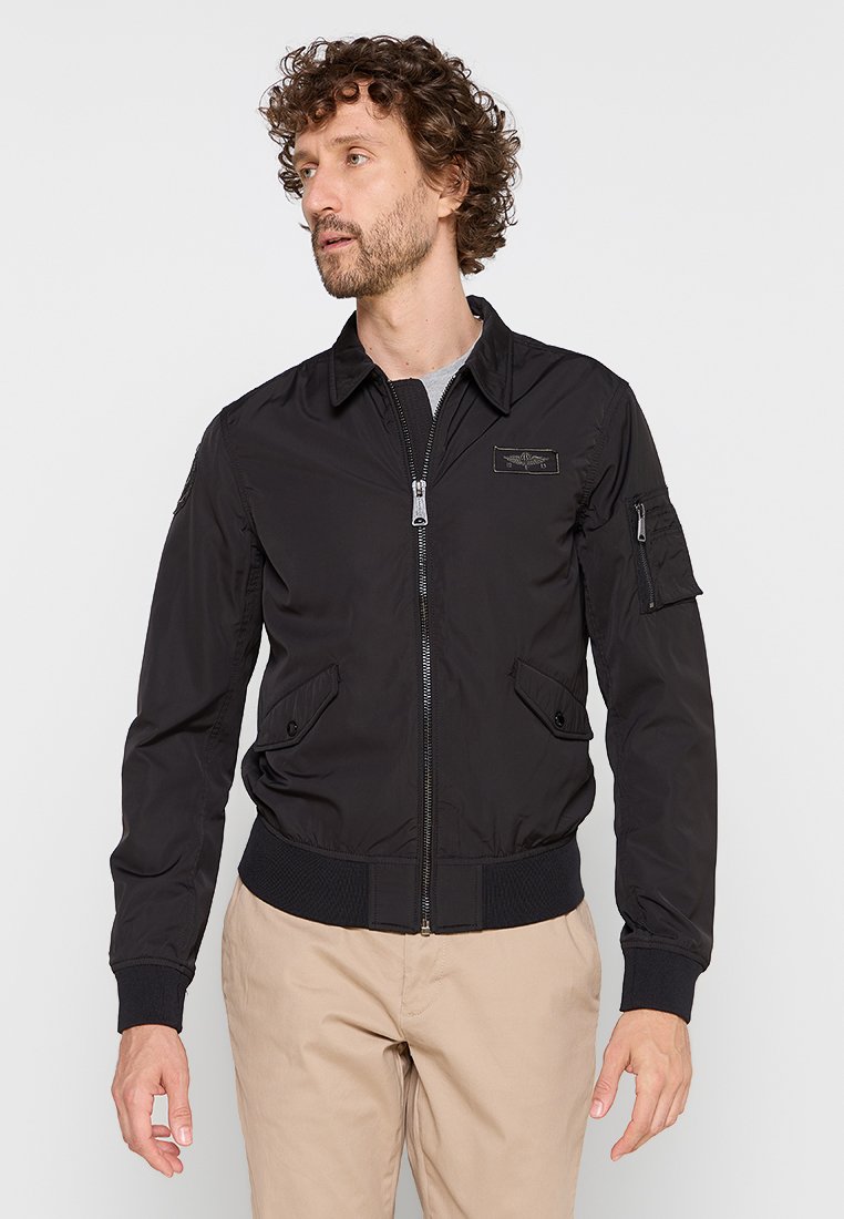 Veste bomber noire en tissu léger avec fermeture éclair à l'avant, poignets et ourlet côtelés, dotée de poches latérales et d'une poche sur la manche.