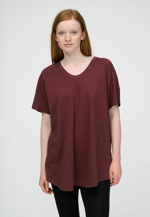 Jonge vrouw met stijl rood haar draagt een losse donkerrode korte mouw T-shirt en een zwarte broek tegen een effen lichte achtergrond.