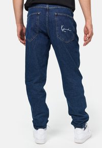 Mörkblå denimbyxor med en avslappnad passform, bakfickor och vit broderad branding. I kombination med vita sneakers, som visar en avslappnad stil.