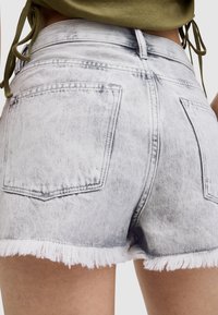 Bermudas de mezclilla en gris claro con un dobladillo deshilachado, que cuentan con dos bolsillos traseros y detalles de costura en contraste. Tejido texturizado con acabado desvanecido.