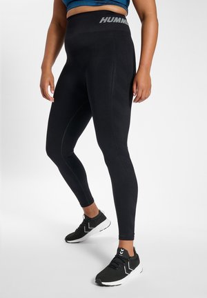 Schwarze Sportleggings mit hohem Bund, strukturiertem Muster und reflektierendem Logo. Getragen mit schwarzen Sportschuhen mit weißen Akzenten.