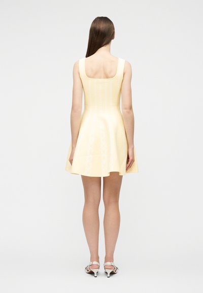 Γυναίκα με μακριά καστανά μαλλιά που φοράει αμάνικο, ανοιχτό κίτρινο φόρεμα με φούστα που ανοίγει και λευκά σανδάλια με τακούνι, στέκεται με την πλάτη προς τα εμπρός σε λευκό φόντο.