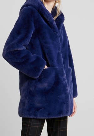 Kvinde iført en marineblå imiteret pelsfrakke med hætte og sidelommer, kombineret med sorte ternede bukser mod en ens grå baggrund.