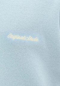 Lys blå sweater med en strikket tekstur, der har "Originals Studio" broderet i cremefarve. Enkel design, ingen yderligere mønstre eller hardware.