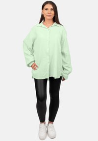 Camicia oversize verde chiaro con superficie testurizzata, abbinata a leggings neri aderenti e sneaker bianche.