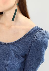 Denim-paitapusero, jossa on pullotetut hihansuut, pyöreä kaula-aukko ja raaka reunus. Vihreä tupsikorvakoru, jossa on kultaiset yksityiskohdat, täydentää asua.