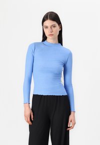 Maglietta a maniche lunghe blu a costine con scollatura e orlo ondulati, abbinata a pantaloni neri plissettati. Il tessuto ha una consistenza liscia.