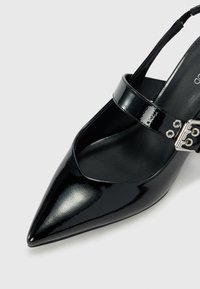 Svart lackläder klackskor med spetsiga tån, tunn ankle strap och en metallisk spänne. Slät textur och glänsande finish.