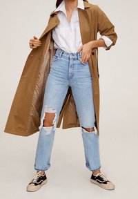 Un abrigo trench marrón sobre una camisa blanca, combinado con unos jeans claros desgastados que presentan grandes rasgaduras y zapatillas negras con una suela crema.