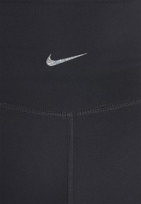 Leggings de sport noirs en tissu lisse. Présentent un logo Nike argenté brillant à la taille. Comprennent des détails de couture visibles.