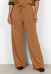 Pantalones marrones de pierna ancha con detalles de costura frontal, cintura con cordón, usados con zapatos puntiagudos marrón oscuro sobre un suelo claro y fondo blanco.