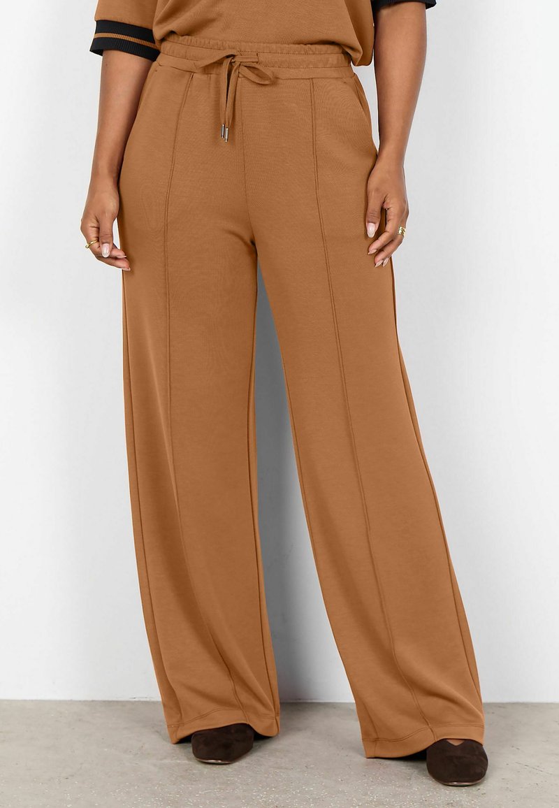 Pantalones marrones de pierna ancha con detalles de costura frontal, cintura con cordón, usados con zapatos puntiagudos marrón oscuro sobre un suelo claro y fondo blanco.