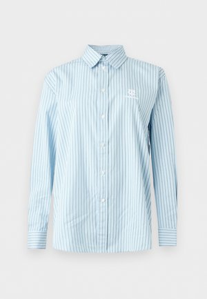 Kvinde med langt mørkt hår iført en blå stribet skjorte, vidde jeans, hvide sneakers og holder en brun læderhåndtaske.