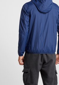 Navy blue lightweight rain jacket with a hood, elastic cuffs, and a cinched hem. Worn over dark gray sweatpants with cargo pockets.
