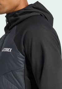 Veste à capuche noire avec un panneau gris foncé brillant et des accents noirs mats, arborant le logo "Terrex" en blanc sur le côté supérieur gauche.