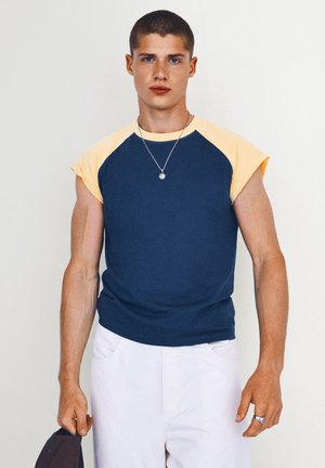 Jeune homme aux cheveux courts portant un débardeur bleu et jaune, un pantalon blanc, un collier en argent, tenant un chapeau de couleur foncée dans une main.