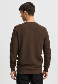 Brun strikket genser med en teksturert mønster, lange ermer, og ribbestrikket halslinning og kant. Modellen vises fra baksiden.