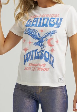 Kobieta w białej koszulce z nadrukiem niebieskiego orła, różowych gwiazdek oraz napisem "Gailey Wilson Shooting for the Moon", w połączeniu z błyszczącymi dżinsowymi spodniami.