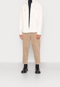 Witte katoenen shirtjacket met drukknoopjes, gecombineerd met tan kleurige corduroy broek en zwarte veterschoenen, waarmee een casual gelaagde look wordt getoond.