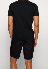 Homme portant un t-shirt noir uni à manches courtes et un short noir au genou, vu de dos sur un fond uni.