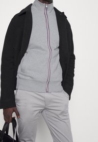 Chaqueta exterior negra sobre un sudadera gris con cremallera y cuello alto, que presenta una franja multicolor en la cremallera, combinada con pantalones gris claro.