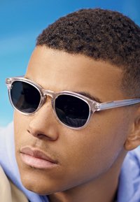 Gafas de sol con marco transparente y lentes negros, de forma redonda y con un sutil patrón en las patillas. Vista en primer plano con un fondo azul.