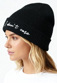 Zwarte gebreide muts met een geribbelde textuur en witte geborduurde tekst "I don't care" op de rand, schuin gedragen.