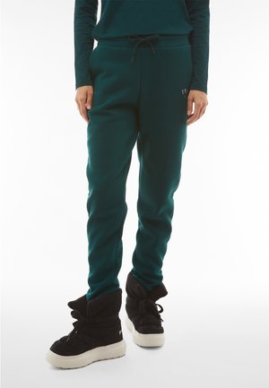 Osoba w ciemnozielonych spodniach dresowych z elastycznym pasem i czarnych wysokich sneakersach z białą podeszwą, stojąca na białym tle.