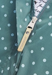 Turkusowa kurtka w białe grochy, z srebrnym zamkiem błyskawicznym i beżowym uchwytem do suwaka z wytłoczonym logo. Wnętrze wykonane z tkaniny we wzory.