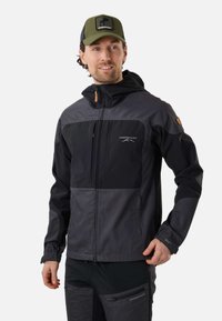 Homme portant une veste et un pantalon d'extérieur gris foncé et noir avec des logos Swedemount, et une casquette Swedemount vert olive, souriant légèrement.